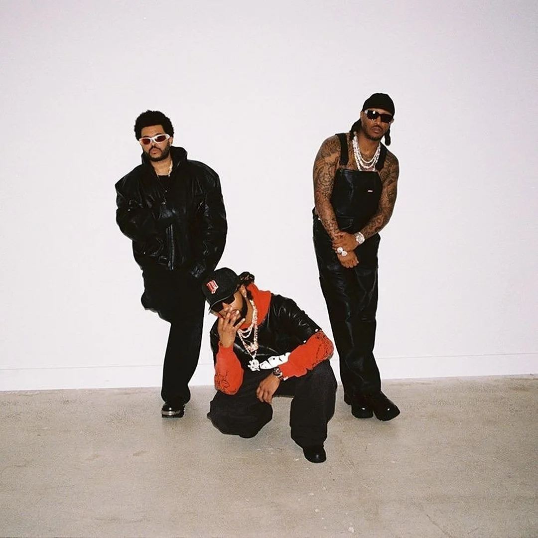 Three men posing against a plain white studio backdrop wearing black leather jackets, overalls, sunglasses, and layered chains