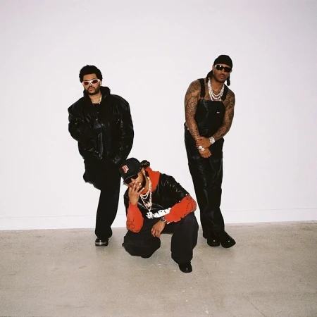 Three men posing against a plain white studio backdrop wearing black leather jackets, overalls, sunglasses, and layered chains