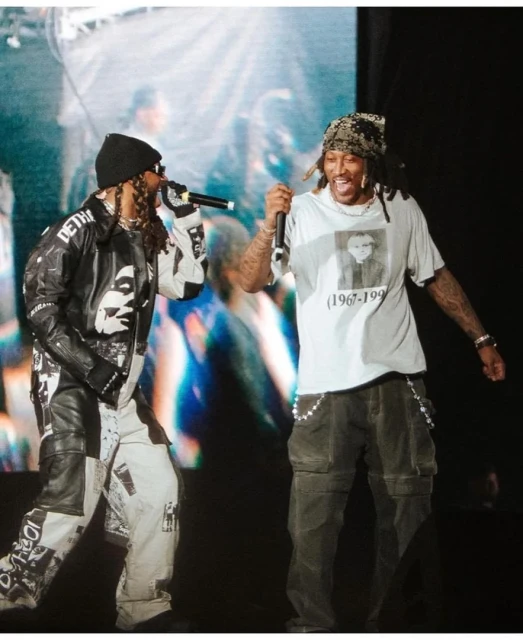 Two adult male rappers perform on stage, one in a graphic jacket and the other in a t-shirt, singing into microphones.