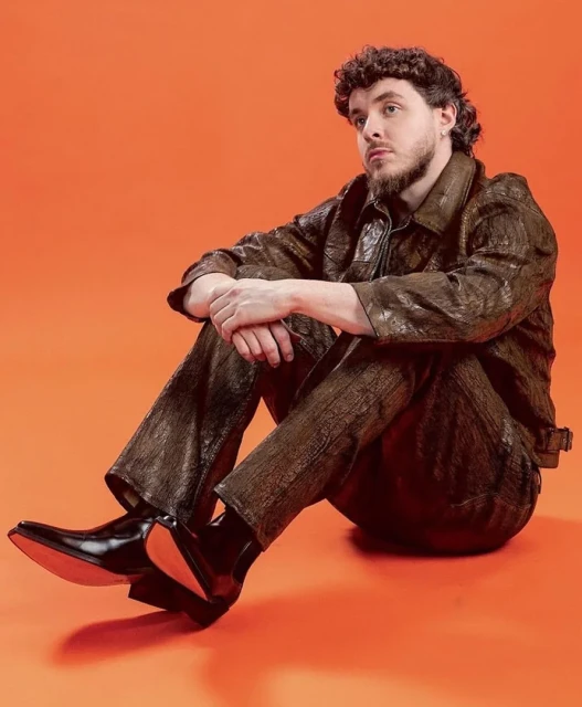 Young man with curly hair and a goatee poses while sitting in a brown reptile-patterned jacket and pants against an orange background.