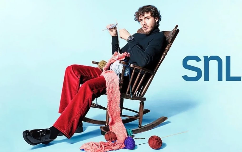 Man in a rocking chair knitting a pink scarf with knitting needles and yarn, with the SNL logo in the background. 
