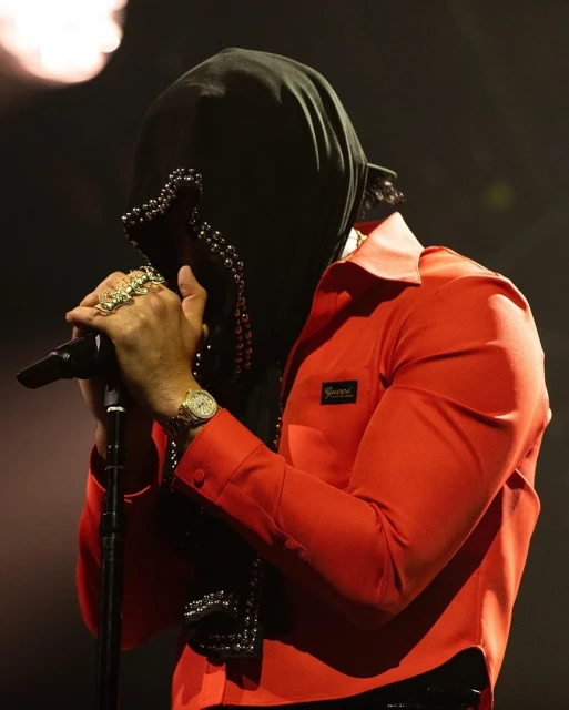 Male singer in red shirt and black hood performs on stage, holding microphone, wearing jewelry and watch in a dimly lit setting.