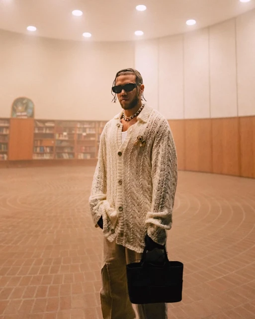 An adult male stands in a large, empty room with books in the background, wearing sunglasses, a cardigan, and holding a black tote bag.
