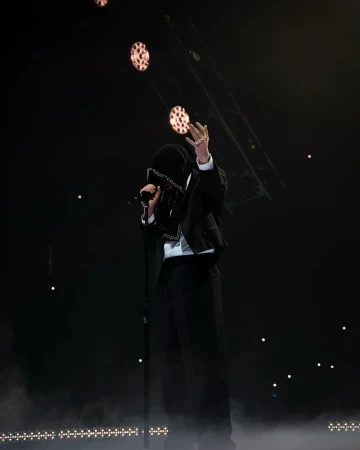 Performer in a black hooded suit and white shirt singing into a microphone on a dark stage with atmospheric floor smoke and overhead theatrical lights.