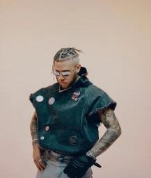 Man with braided hair wearing a dark distressed sleeveless hoodie adorned with pins, styled by Storm Pablo.