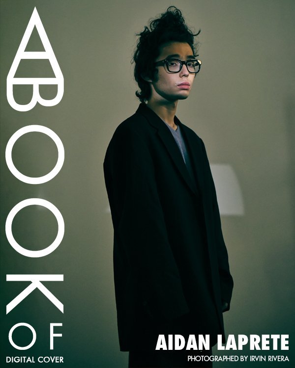 Portrait of a young adult with dark hair and glasses wearing a black blazer in a photo studio with minimalist lighting.