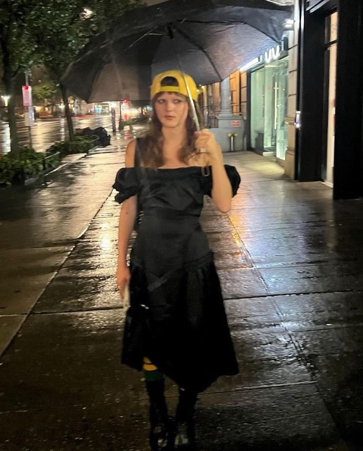 Woman in a black dress and yellow cap walking on a wet city street at night under an umbrella, streetlights reflecting off the pavement.