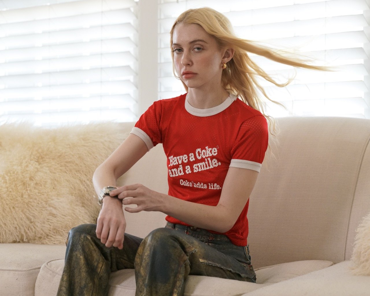 Young woman in red t-shirt with gold pants sitting on a white couch in a brightly lit indoor setting with white window blinds.