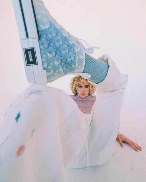 Fashion shot of a woman with curly hair, wearing a lavender sweater and white pants, with a blue Dior shoe on her leg.