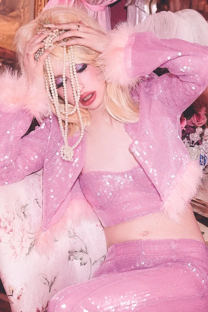 Woman in sequin pink jacket and pants poses with pearls and tiara against a soft floral backdrop for a fashion editorial shot.