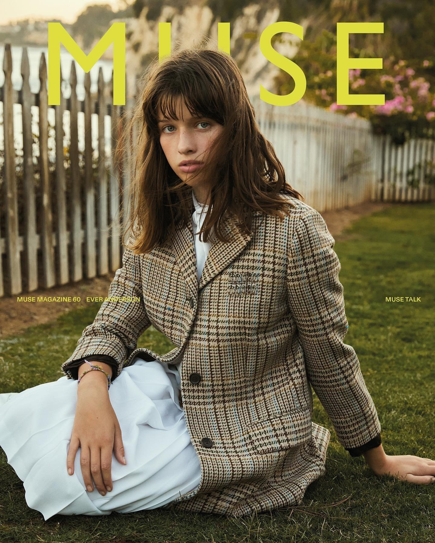 Magazine cover featuring a young woman in a tan plaid blazer and white dress, sitting on grass with trees and a fence in background.