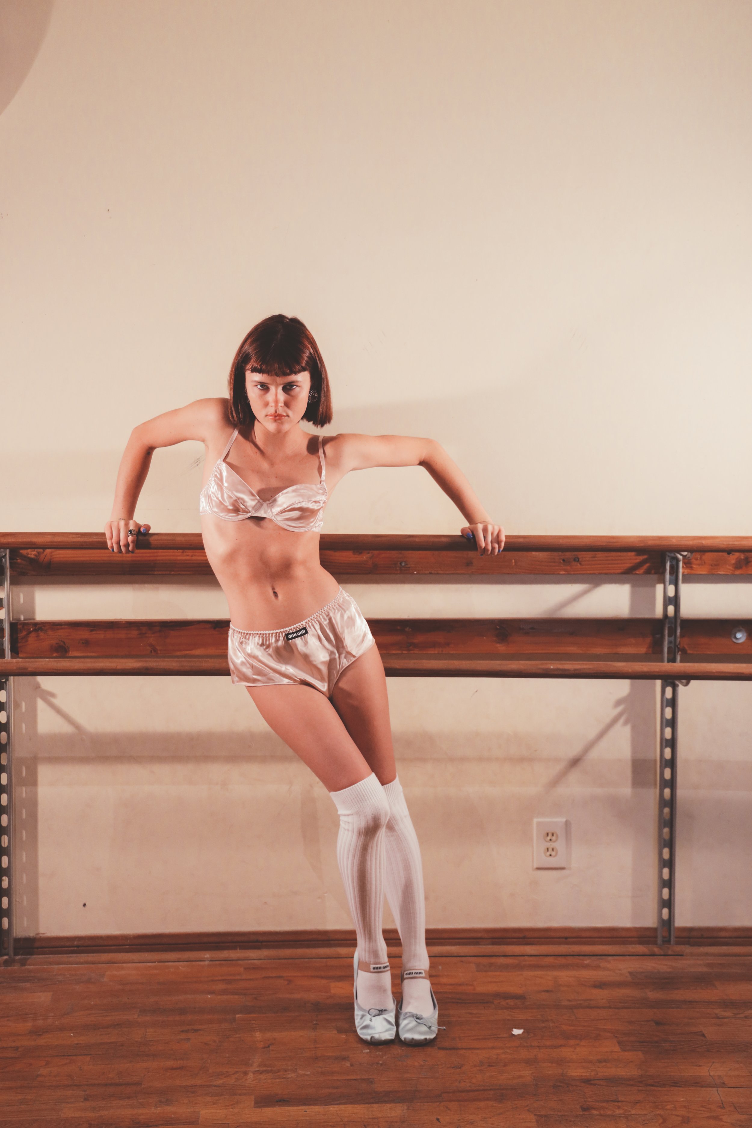 A woman in lingerie poses leaning against a wooden ballet barre in an indoor setting with a wooden floor and neutral background.