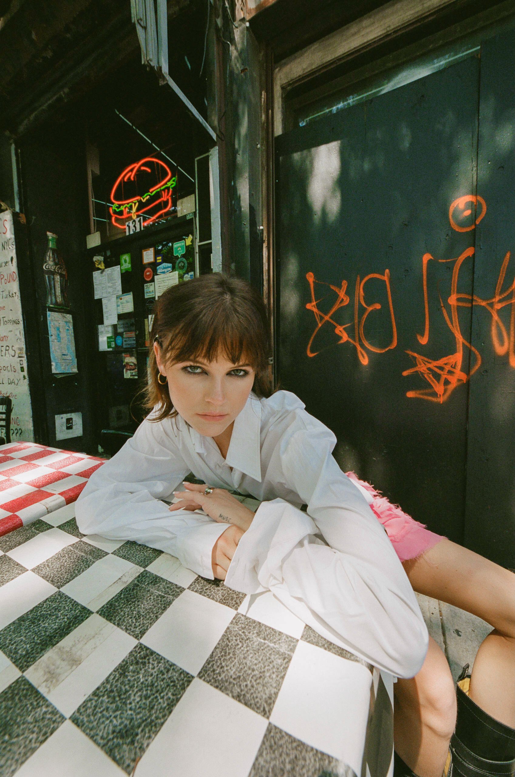 Brunette woman in a white shirt sits at a checkered table, with neon burger sign, graffiti background, and natural light.