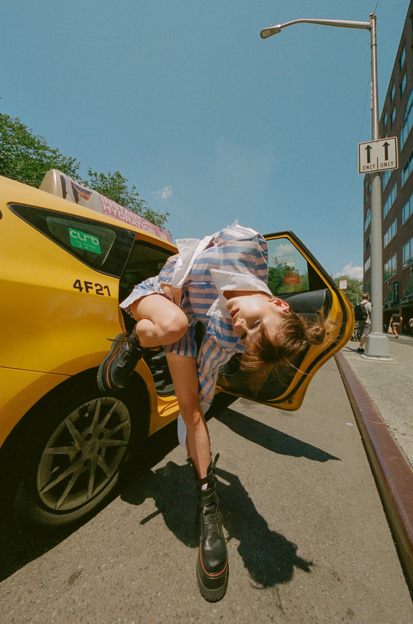 Young woman leans out of a yellow taxi on a city street on a sunny day, wearing a striped top, black boots, and a playful pose.