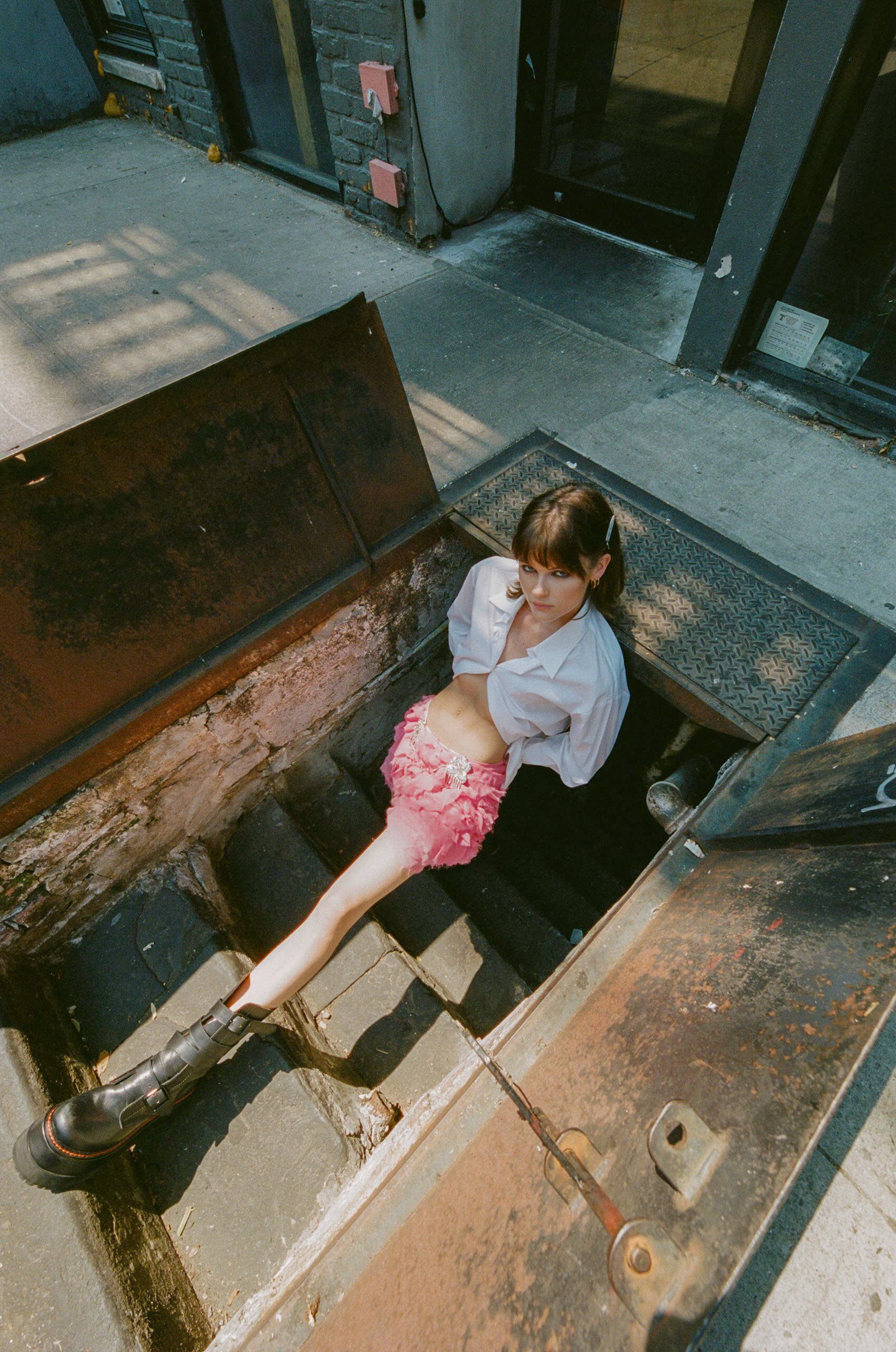 Young adult woman in a white shirt and pink skirt sits on concrete stairs leading down to a dark space on a sunny city street.