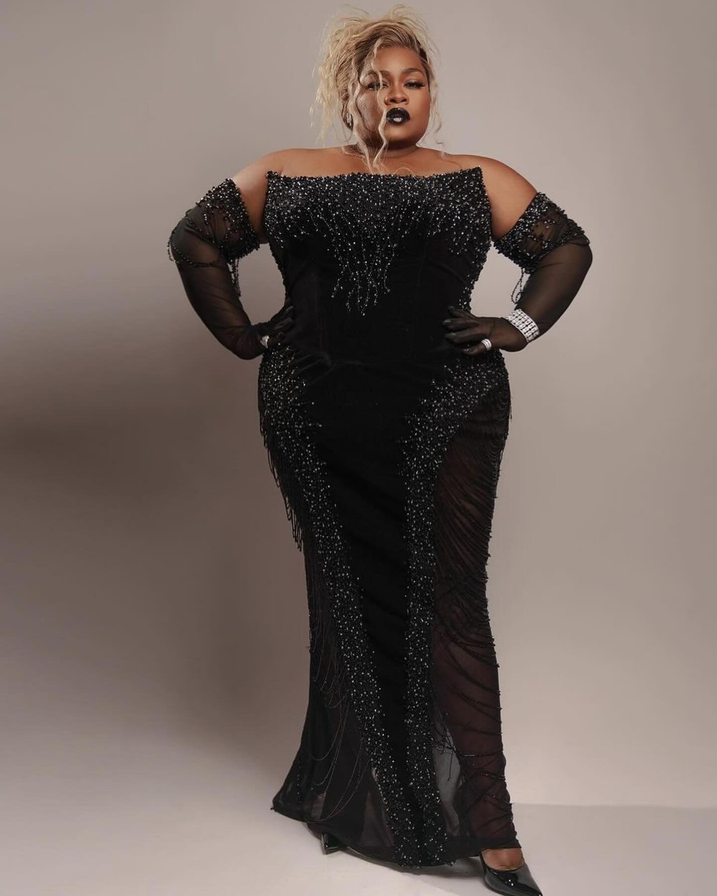 Full-length portrait of a plus-size woman in a black off-the-shoulder gown with sheer sleeves posing against a light grey backdrop.