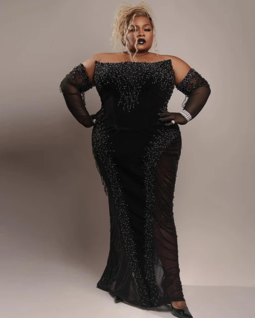 Full-length portrait of a plus-size woman in a black off-the-shoulder gown with sheer sleeves posing against a light grey backdrop.