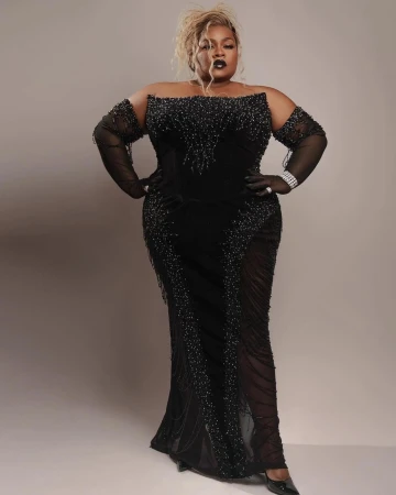 Full-length portrait of a plus-size woman in a black off-the-shoulder gown with sheer sleeves posing against a light grey backdrop.