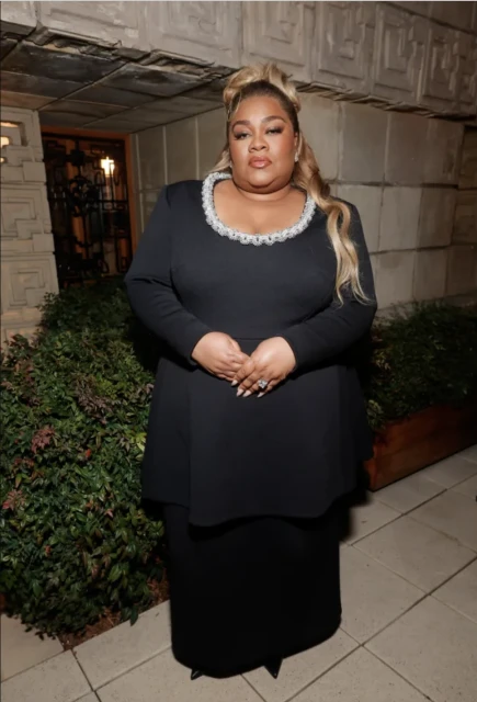 Elegant full portrait of a plus-size Black woman in a long-sleeved black dress with blonde highlights standing indoors.