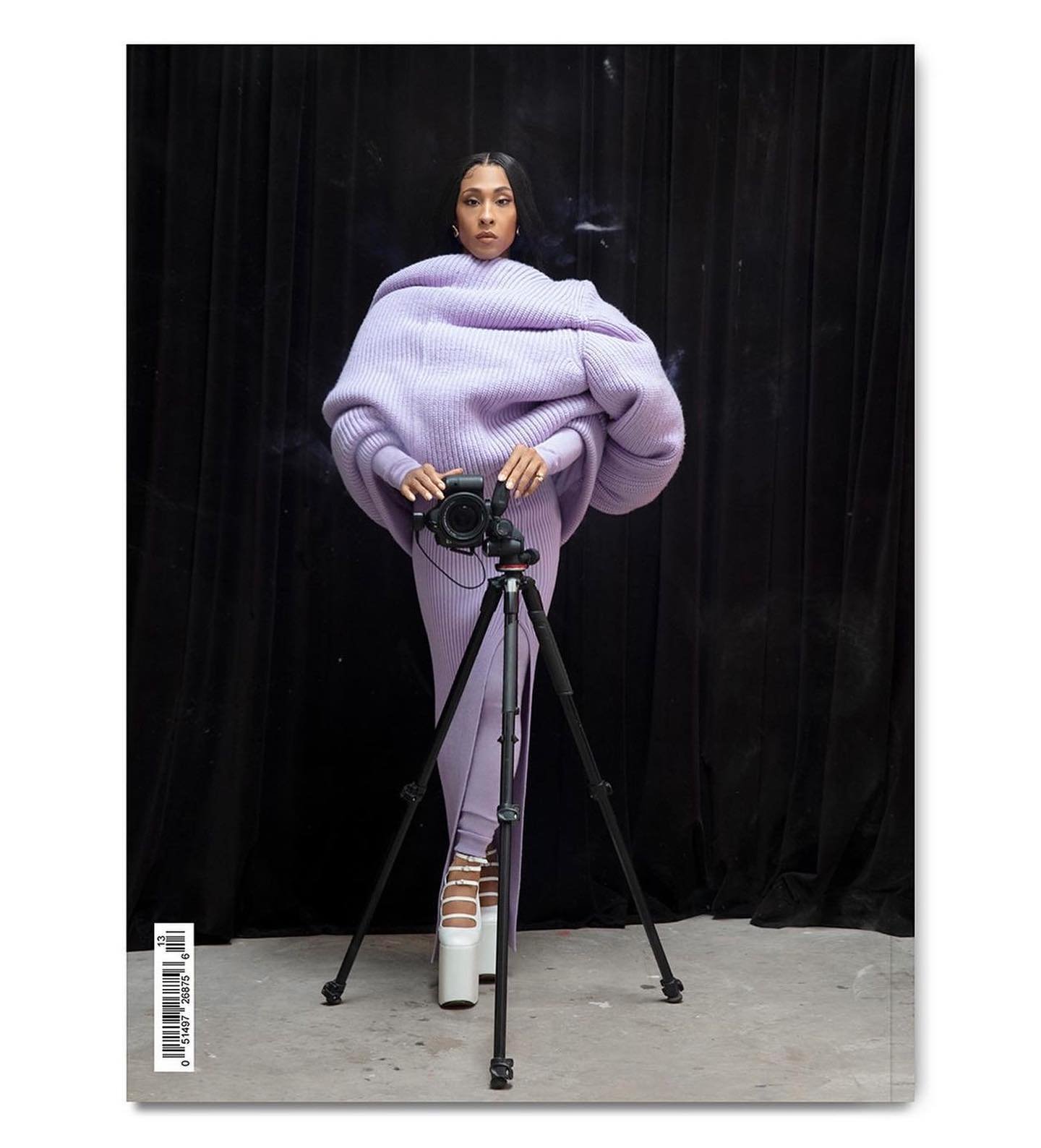 Portrait of a woman in lilac knitwear holding a camera on a tripod, standing in front of a black curtain in a photo studio.