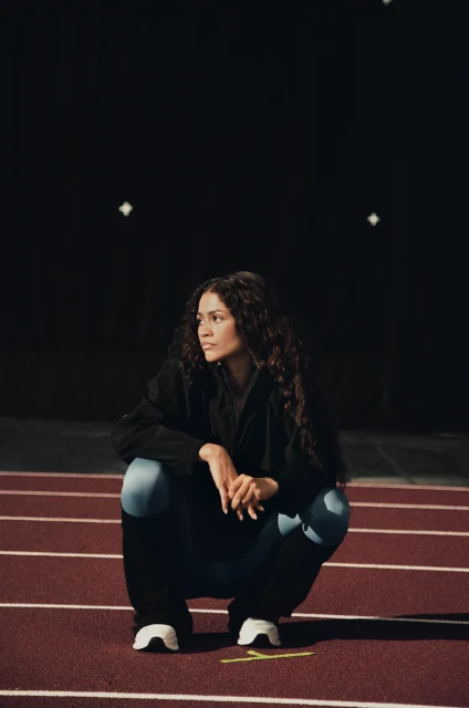 Female athlete with curly hair wearing a jacket and leggings, crouches on a red running track in an outdoor environment at night.