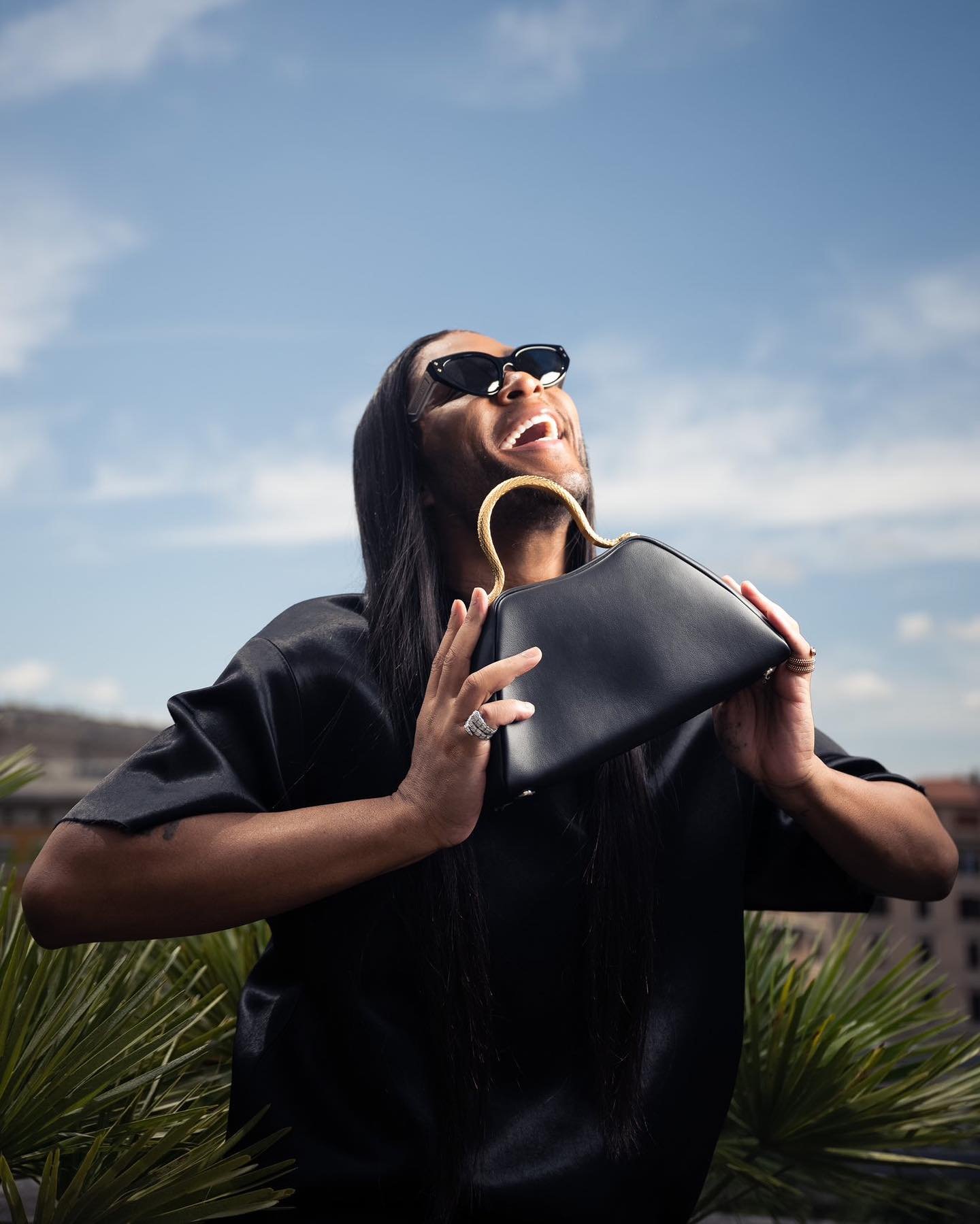 Adult male laughs in the sun while holding a black structured handbag with a gold handle. He's wearing sunglasses and a dark shirt.