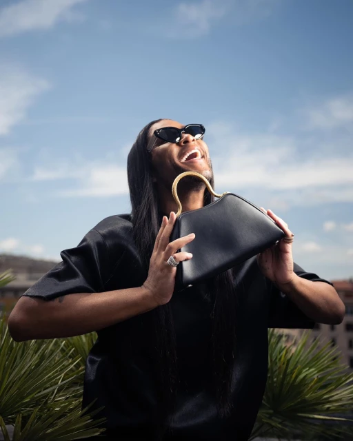 Adult male laughs in the sun while holding a black structured handbag with a gold handle. He's wearing sunglasses and a dark shirt.