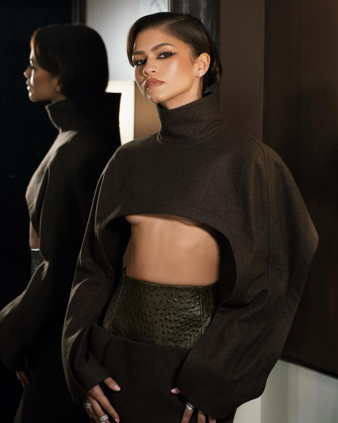 Zendaya poses in studio lighting, showcasing a modern crop top and skirt combination, with a reflected image emphasizing depth.