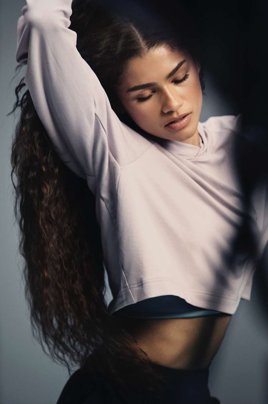 Portrait of a woman with long curly hair wearing a lavender hoodie with a neutral background, showcasing fitness or beauty.