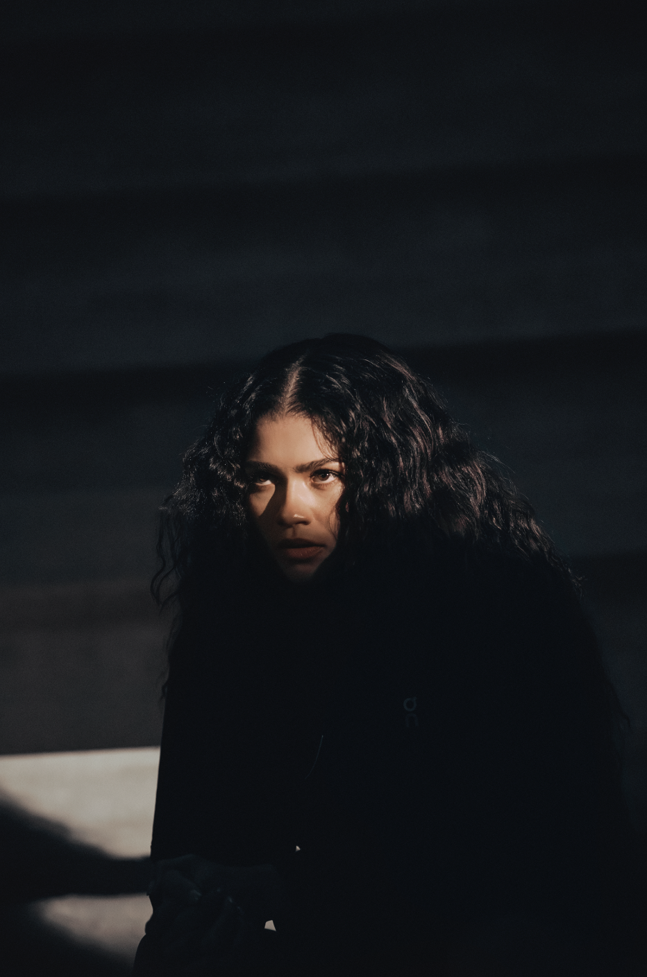 Close-up portrait of a woman with long, dark curly hair and dark clothing, looking directly at the camera in a dimly lit environment.