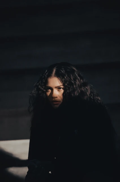 Close-up portrait of a woman with long, dark curly hair and dark clothing, looking directly at the camera in a dimly lit environment.