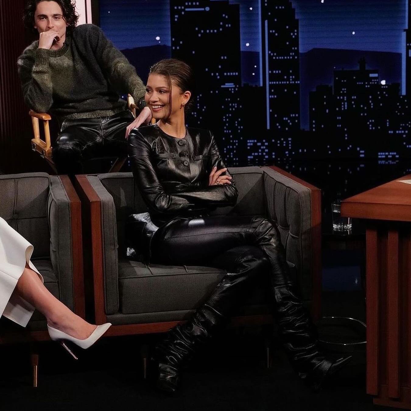 Zendaya sitting on a couch in a black leather outfit with a man in the background, in front of a cityscape backdrop on a late night show.
