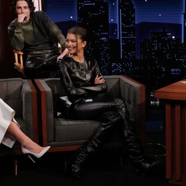 Zendaya sitting on a couch in a black leather outfit with a man in the background, in front of a cityscape backdrop on a late night show.