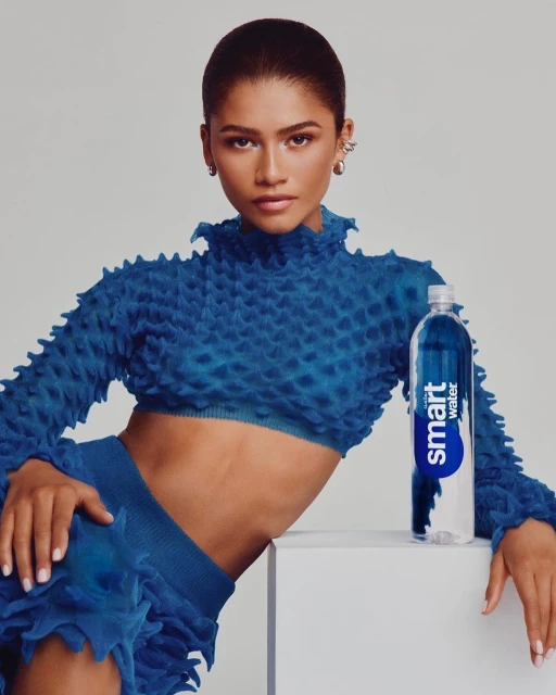 Zendaya poses in a vibrant blue textured outfit next to a Smartwater bottle in a studio setting. Fashion and beauty converge.
