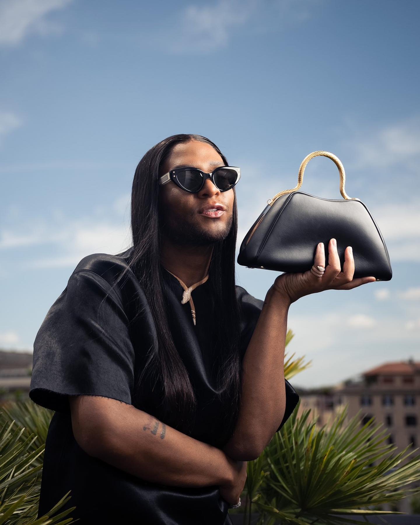 Adult male with long hair poses outdoors holding a black handbag with a gold handle, wearing sunglasses, a necklace, and a black satin shirt.