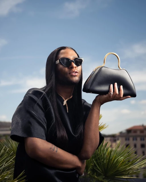Adult male with long hair poses outdoors holding a black handbag with a gold handle, wearing sunglasses, a necklace, and a black satin shirt.