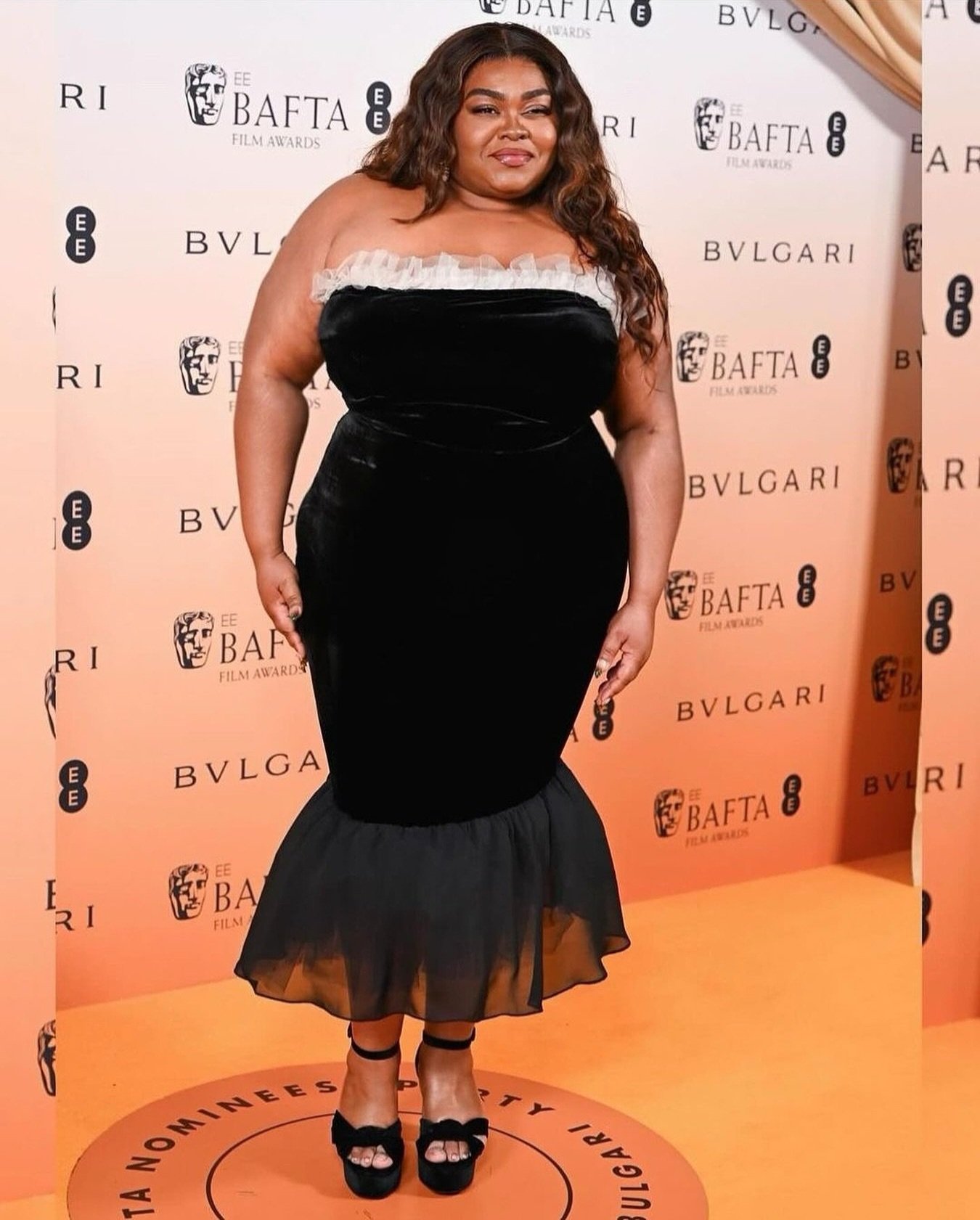 Woman in black strapless dress with black heels poses on orange carpet at film awards show, smiling with long brown wavy hair.