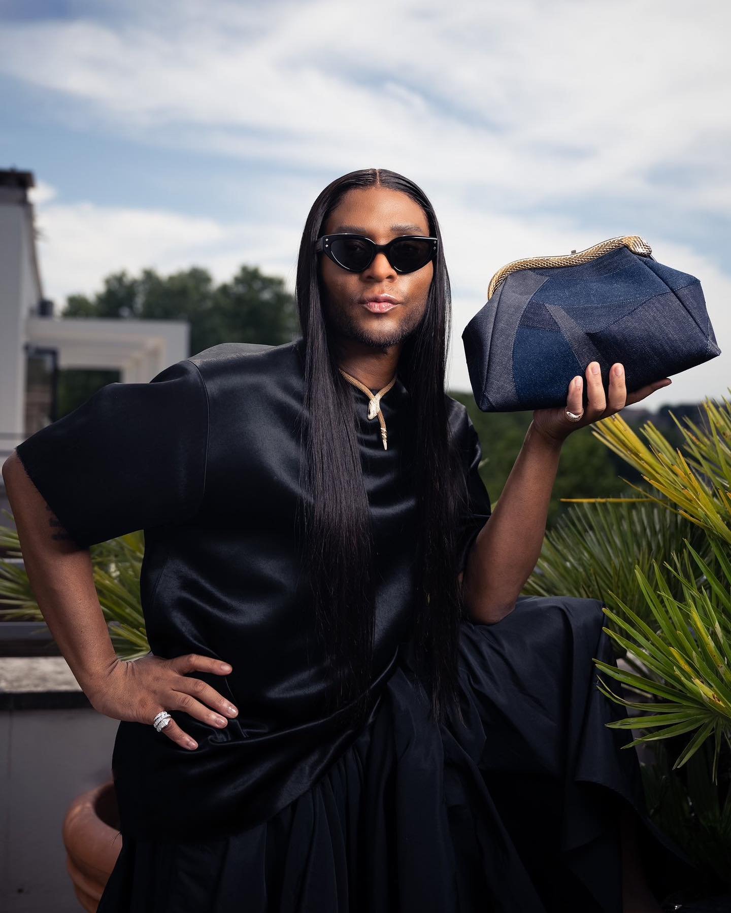 Person in black silk outfit poses with a blue denim clutch outdoors against a sunny sky and architectural background.