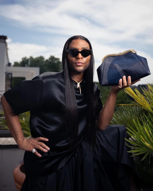 Person in black silk outfit poses with a blue denim clutch outdoors against a sunny sky and architectural background.