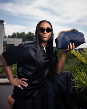 Person in black silk outfit poses with a blue denim clutch outdoors against a sunny sky and architectural background.
