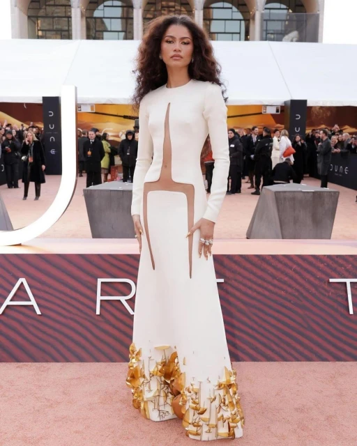 Zendaya poses on a red carpet in a white gown with gold embellishments at the 'Dune' premiere, surrounded by photographers and onlookers.