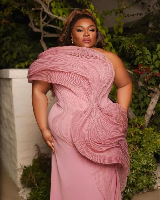 Portrait of a plus-size African-American woman in an elegant pink gown posing outdoors by a white wall with green foliage.