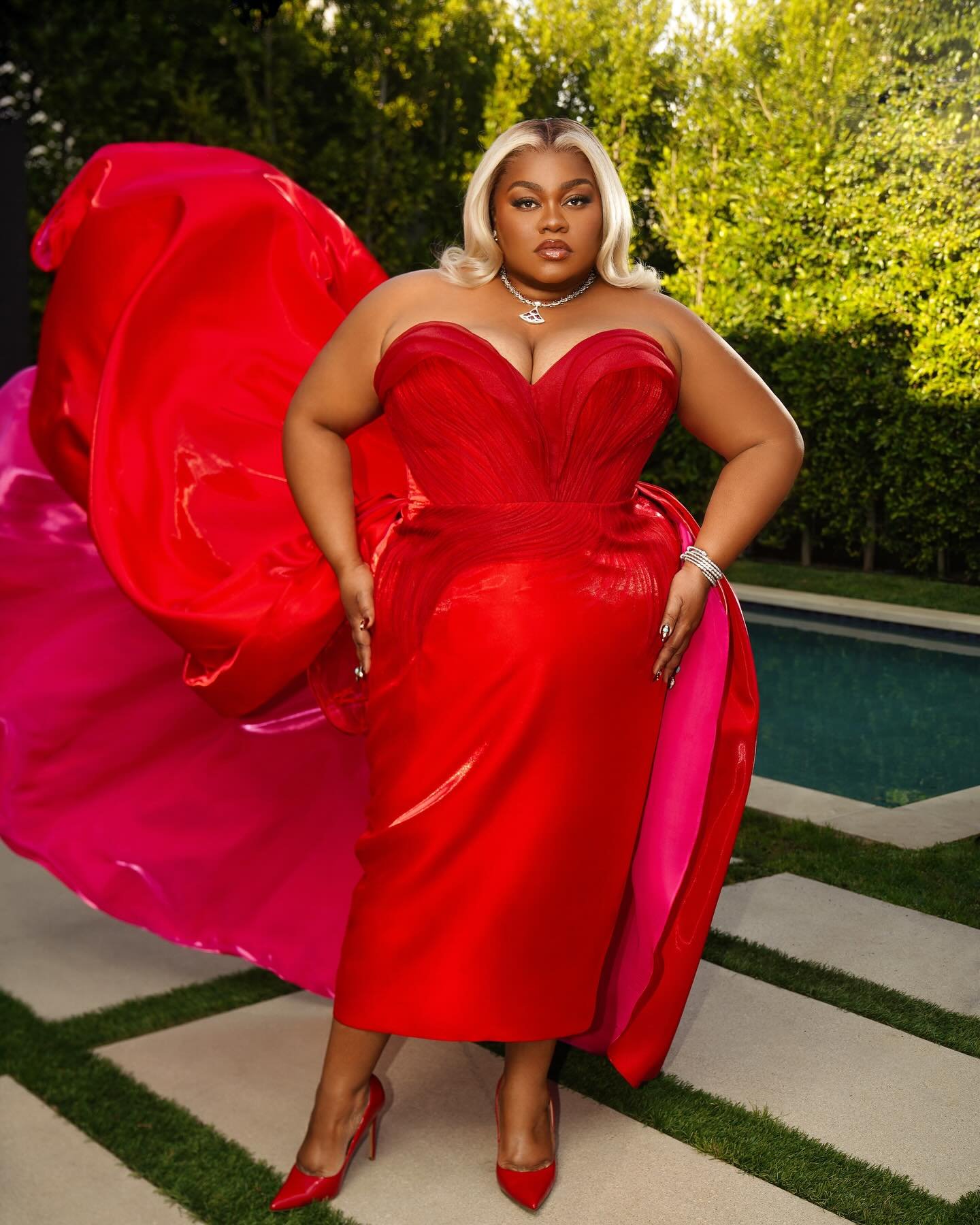Woman with platinum blonde hair posing in a red strapless gown in front of greenery and a light stone tiled patio.
