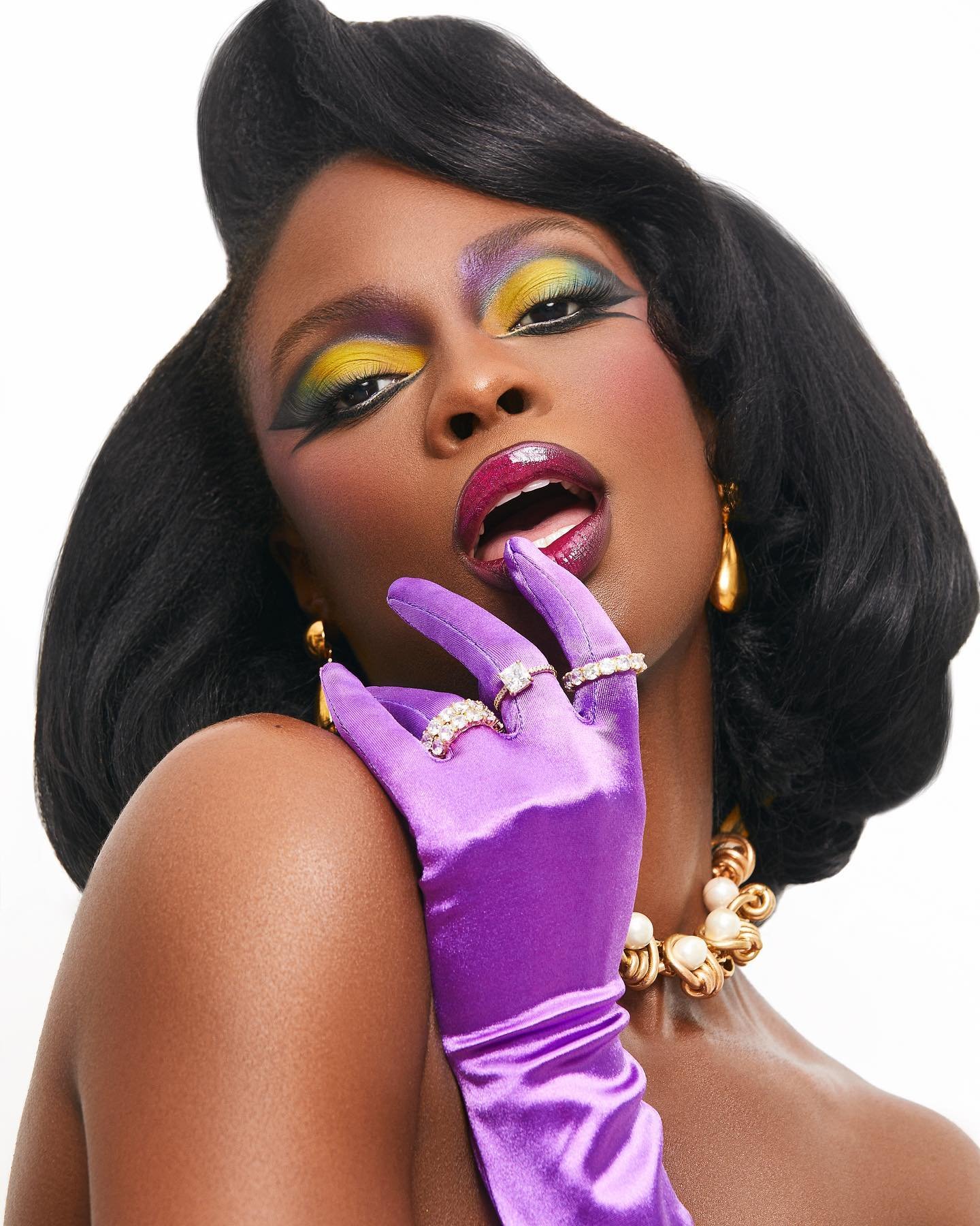 Portrait of a Black woman with dramatic eye makeup, purple satin gloves, and jewelry against a clean white backdrop in a studio setting.