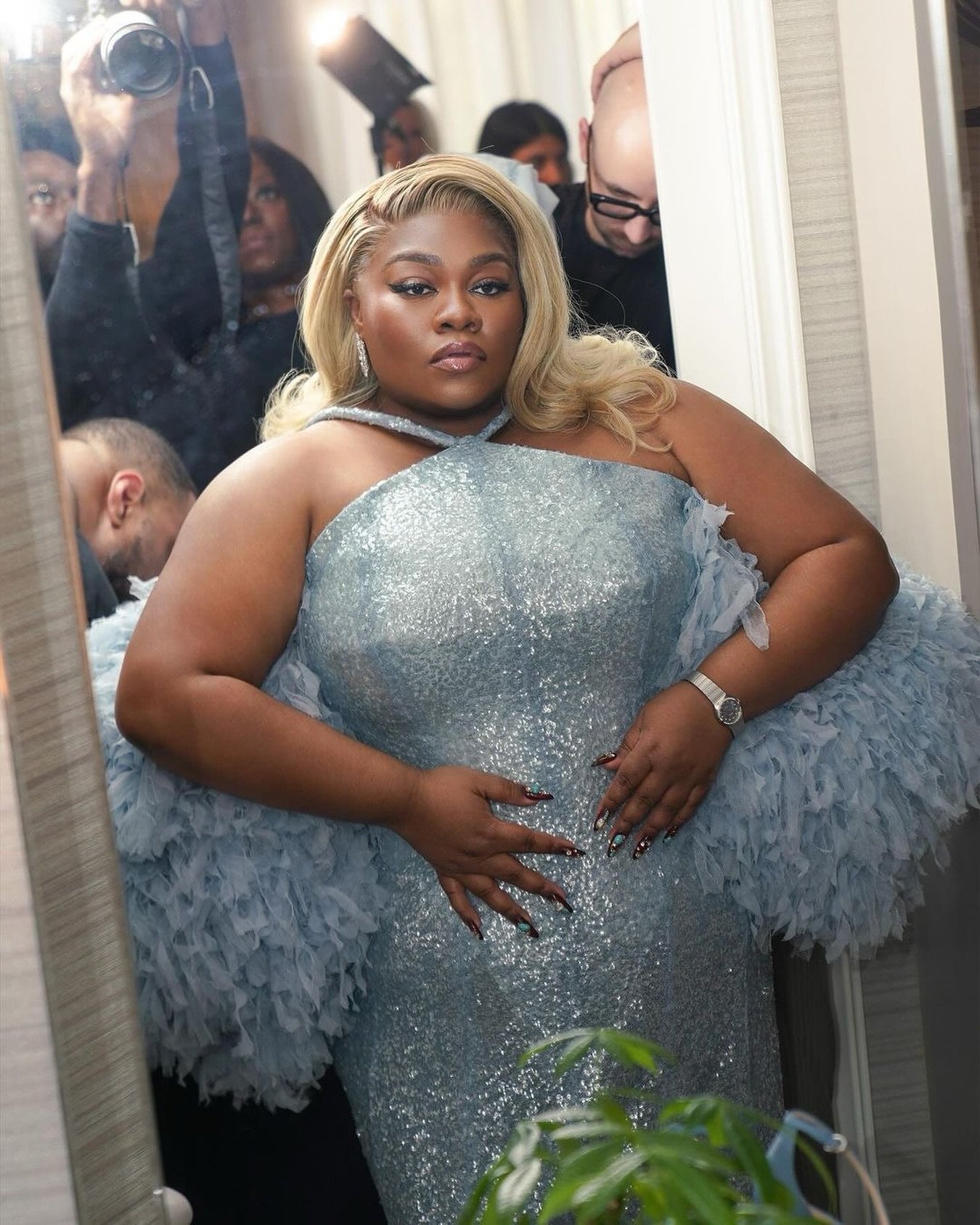 Plus-size Black woman in a sparkling light blue gown with voluminous feathered sleeves surrounded by photographers.