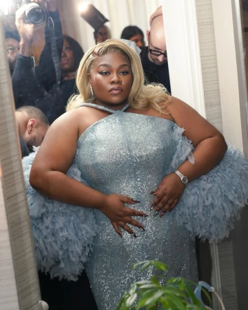 Plus-size Black woman in a sparkling light blue gown with voluminous feathered sleeves surrounded by photographers.