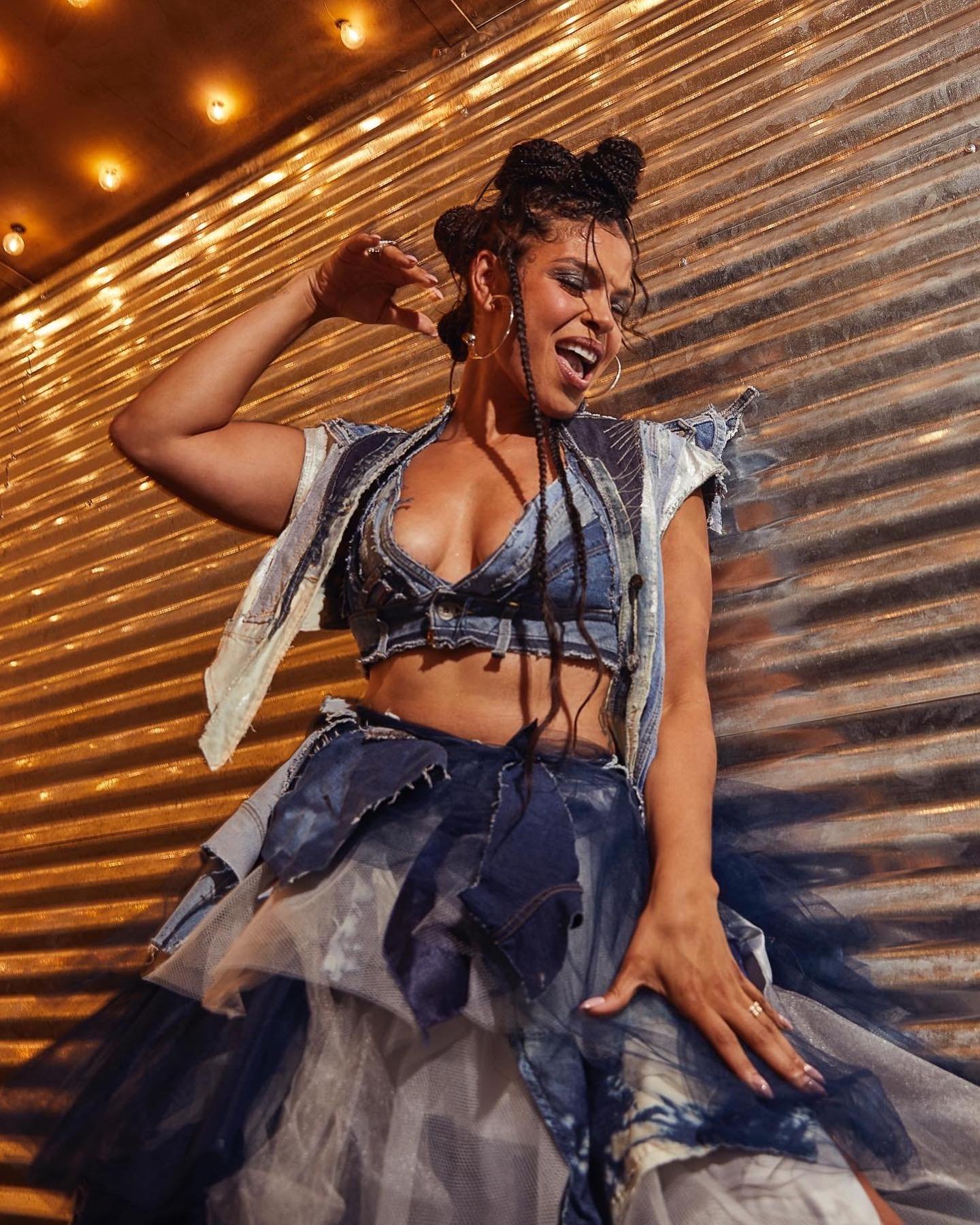 A woman with unique braided hair stands in a denim dress, captured in a medium shot against a corrugated metal wall with warm lighting.