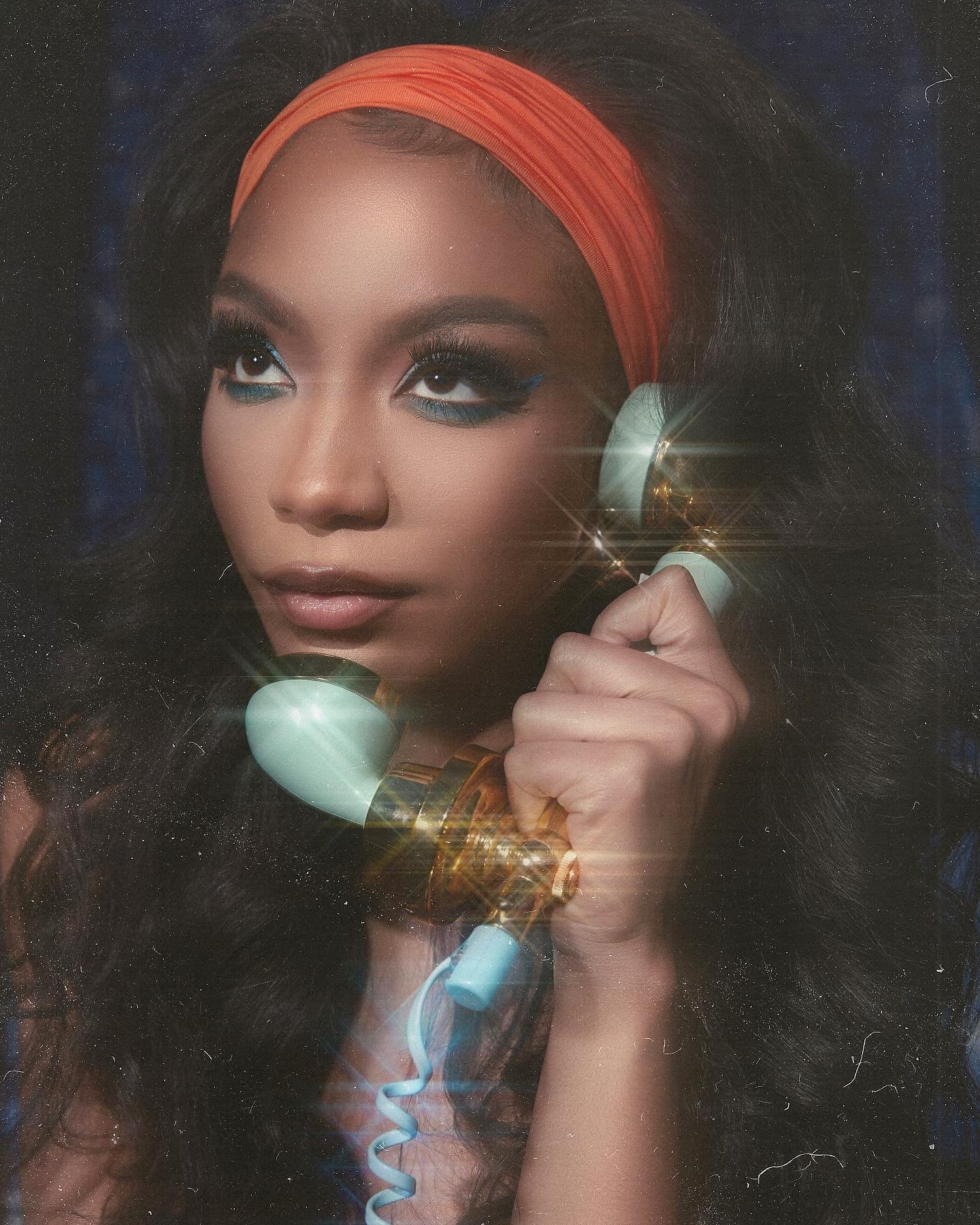 A woman with dark hair and orange headband holds a vintage phone, showcasing makeup and a retro aesthetic against a dark blue backdrop.