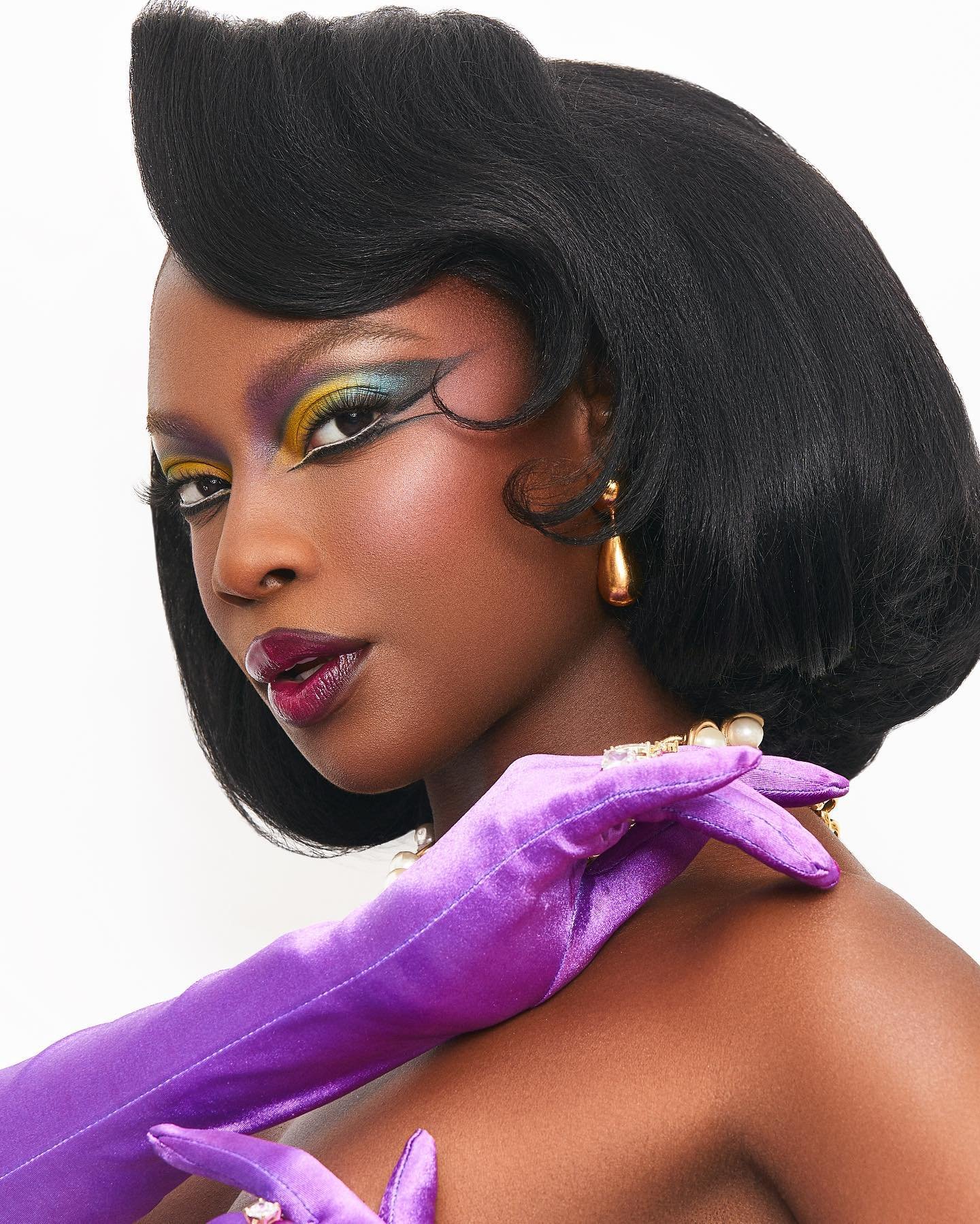 Close-up of a black woman with retro hairstyle, dramatic makeup, gold teardrop earrings, gold necklace, and purple gloves against white.