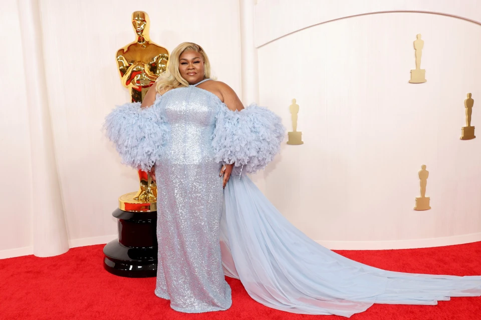 Woman in shimmering light blue gown with feathered sleeves on red carpet in front of a golden Oscar statue and white backdrop.